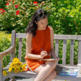 A person in an orange dress sits and paints a watercolor on a sunny day in a garden