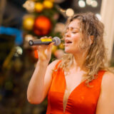 A person with long blonde hair, wearing a red dress, sings into a microphone
