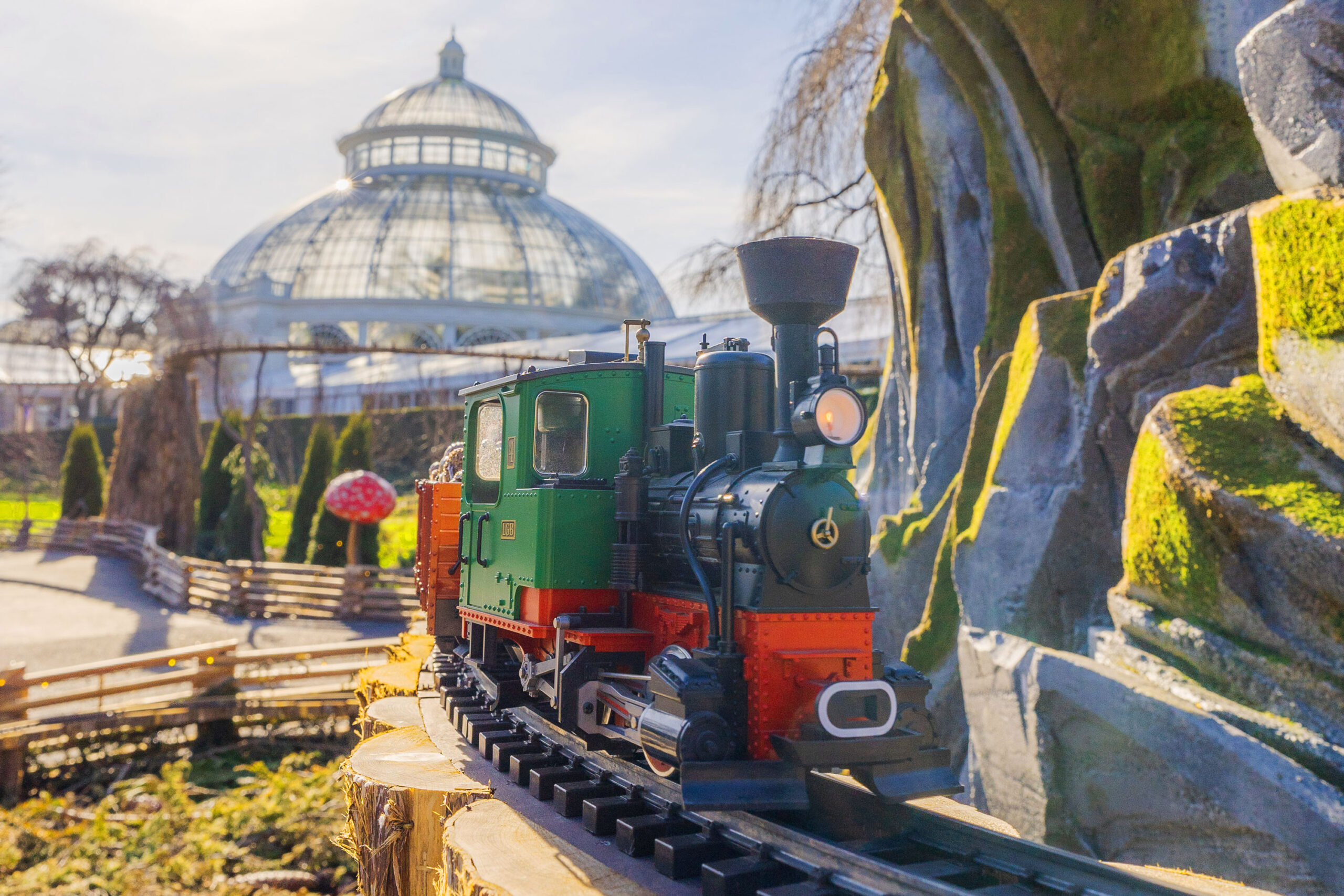 A green and red model train travels along a track outdoors on a sunny day