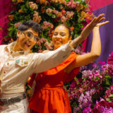 Two people in colorful dance outfits pose for a photo in front of a wall of pink and orange flowers