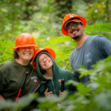 Three people in orange hard hats pose for a photo in a green forest