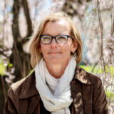 A person in a brown jacket and white scarf poses among sunny trees for a photo
