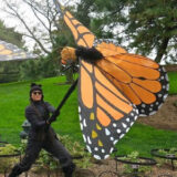 A person in a black outfit holds a large orange butterfly puppet