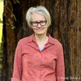 A person with short blonde hair, wearing a glasses and pink shirt, poses for a photo in front of a wide tree trunk