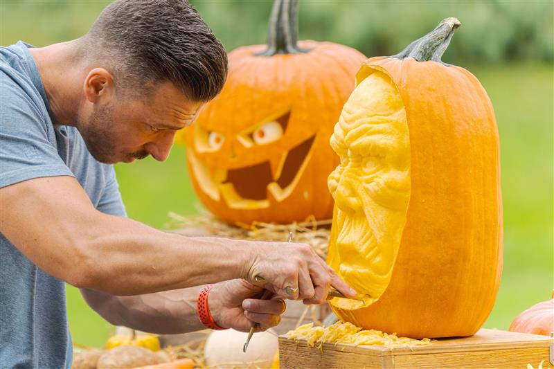 A person with short dark hair uses a tool to carve a face into an orange pumpkin