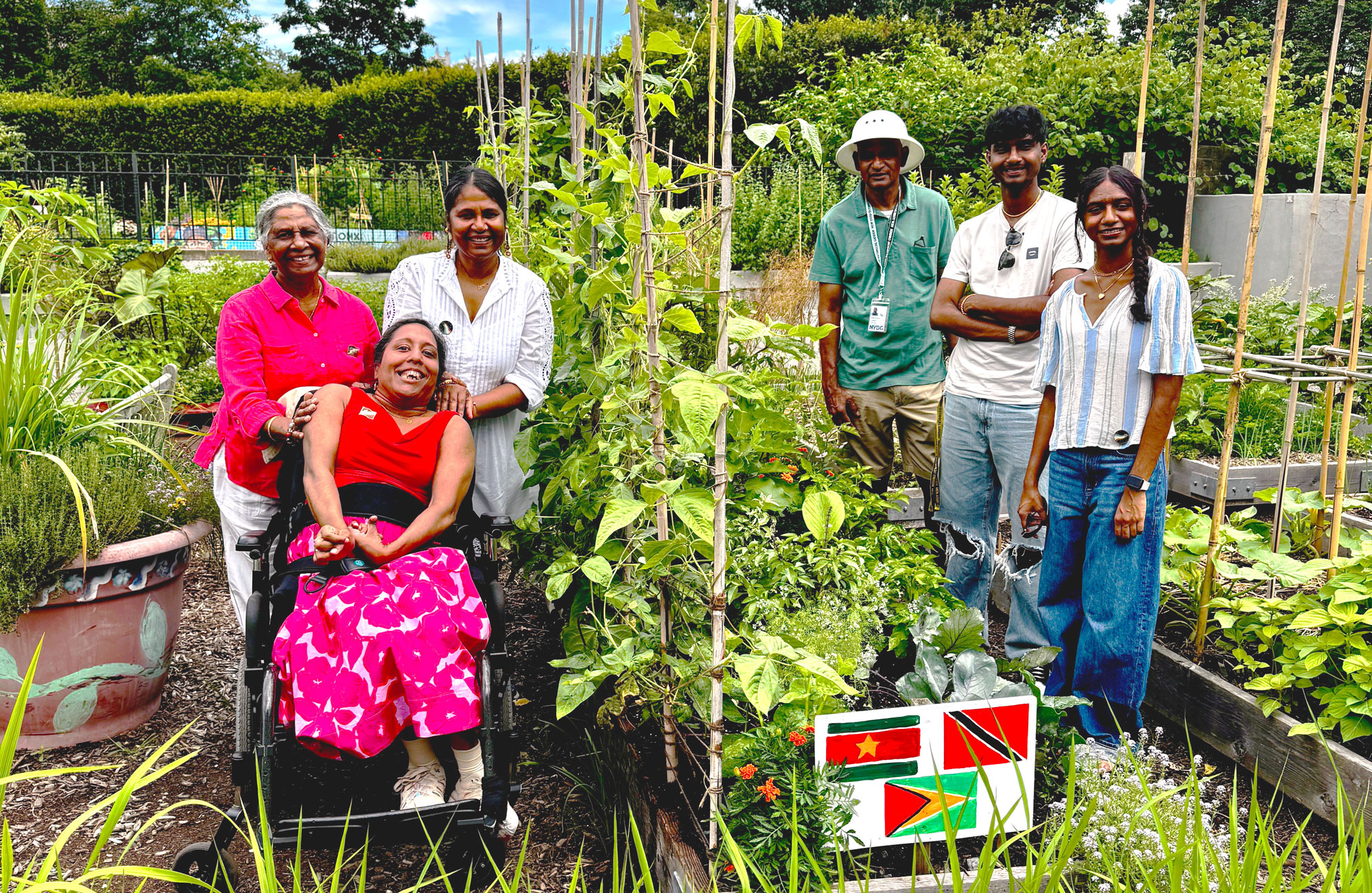 Lakshmee and her joyful multigenerational Indo-Caribbean family which includes her mom, sister, husband and two children smile together among tall, green plants in a thriving garden. Annie sits in a wheelchair wearing a bright pink top and pink floral skirt, surrounded by her family in colorful summer clothes. The group radiates warmth, love, and connection, framed by lush greenery and small flags of Guyana, Suriname, and Trinidad on their garden bed frames representing their Caribbean heritage.