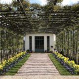A trellis of vines over a flowered pathway leading to a house door