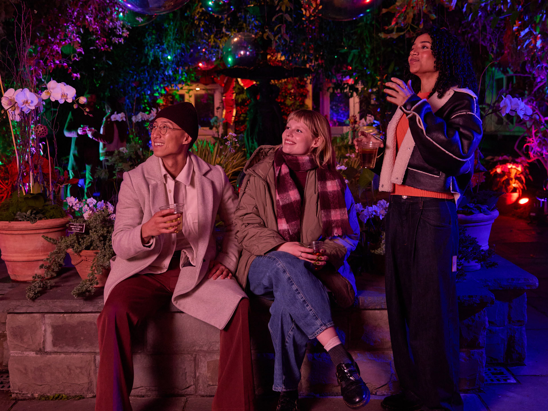 A group of three friends in winter clothes enjoy drinks in a warmly lit holiday atmosphere