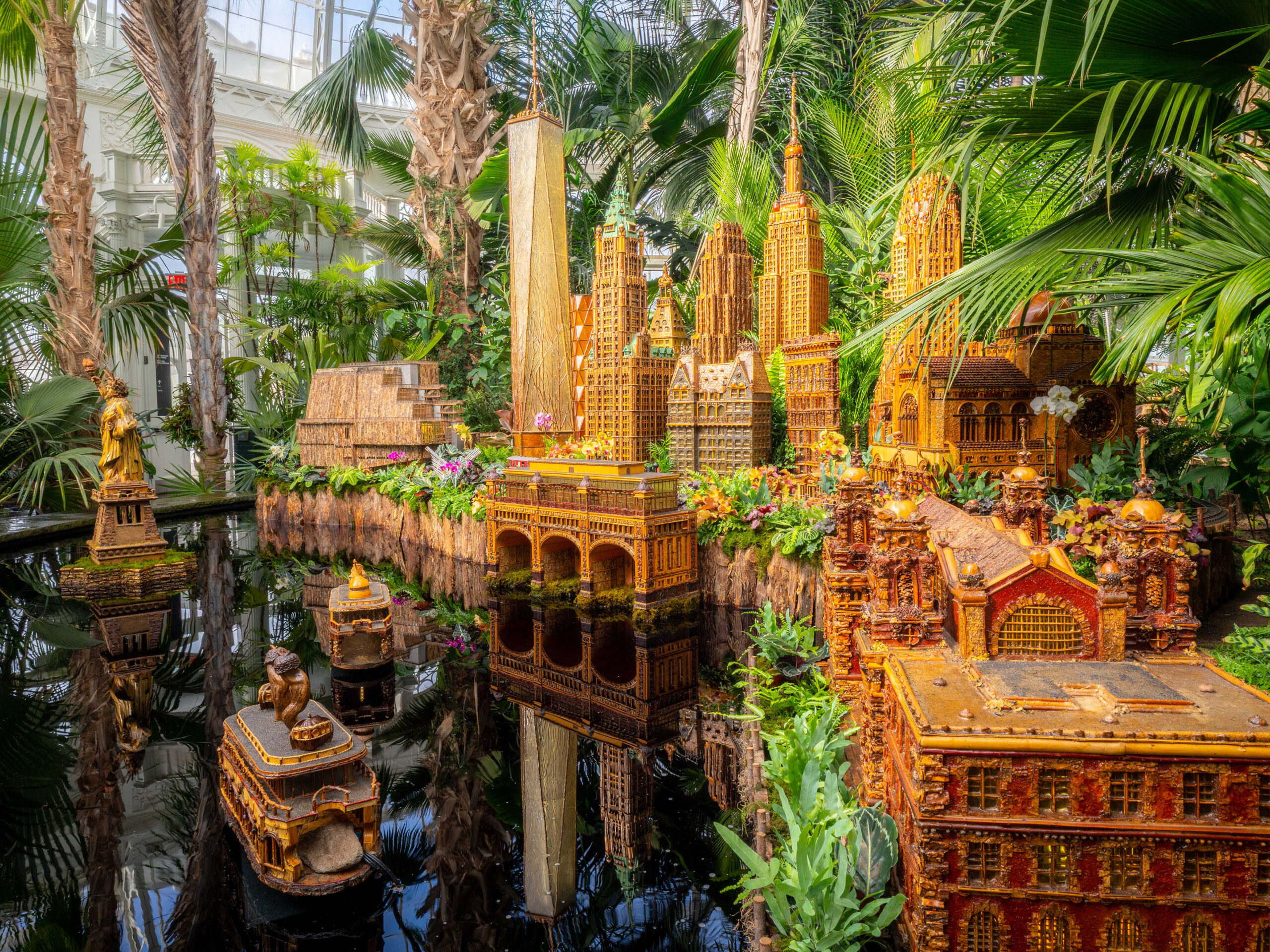 An arrangement of miniature building replicas made of plant parts, set among green foliage and a water feature
