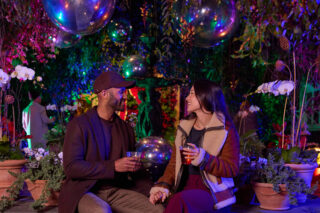 A couple sits under twinkling pink and green holiday lights, smiling and holding drinks.
