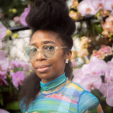 A person in a colorful blue shirt and gold glasses poses for a photo in front of a backdrop of pink flowers