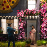 Two people pose for photos among softly lit buildings covered in pink flowers