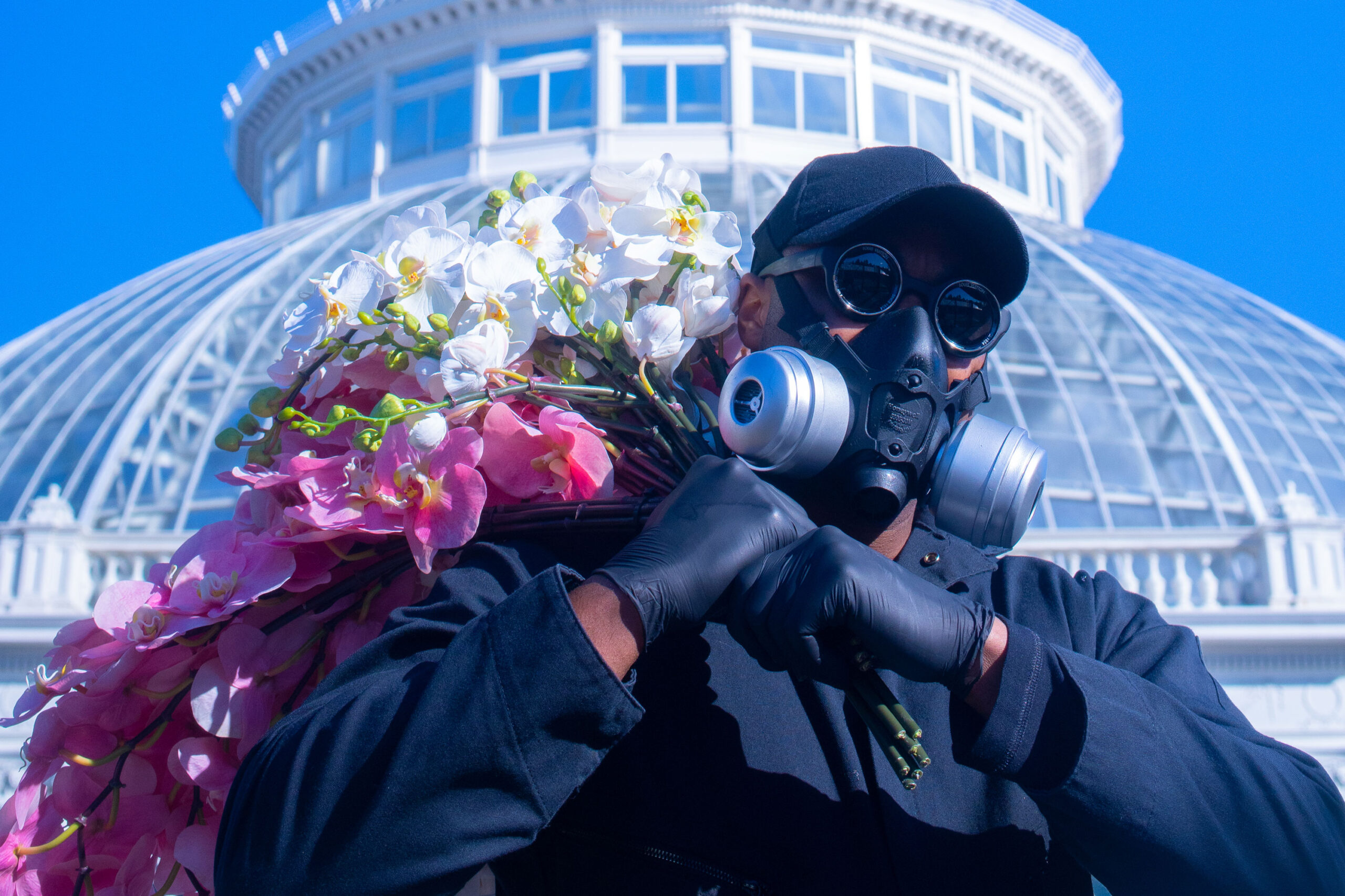 A person in a black outfit, hat, and gas mask hauls a bouquet of colorful pink and white flowers with a white conservatory dome and blue sky visible in the background