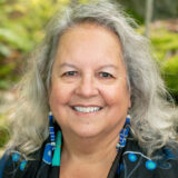 A person with long gray hair, wearing blue earrings, poses for a photo outdoors