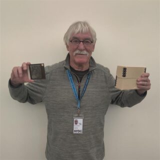 A person with short white hair and a white mustache holds up a glass lantern slide along with a pair of boxes full of additional slides