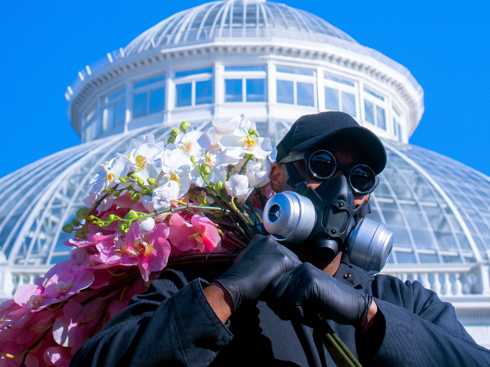 A person in a black hat and mask holds a bundle of pink and white flowers over their shoulder, with a white conservatory dome beneath a blue sky in the background