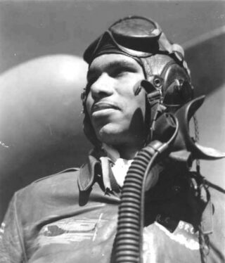 A person in a World War II fighter pilot's outfit poses for a black and white photo in front of a plane