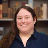A person with long dark hair and a blue shirt poses for a photo in a library