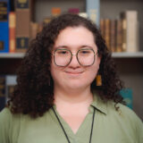 A person with long dark hair, glasses, and a green shirt poses for a photo in a library