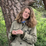 A person with long blonde hair, wearing a tan coat, poses for a photo near a tree