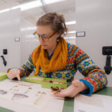 A person in a colorful shirt and orange scarf examines dried plant specimens in a brightly lit archival space