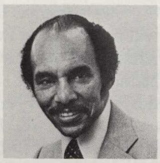 A black and white photo of a person in a gray suit and dark tie, with a mustache