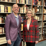 A person in a purple blazer and a person in a red plaid blazer pose for a photo together in a library