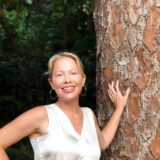 A person with blonde hair, wearing a white vest, leans against a brown tree trunk while being photographed