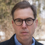 A person with short brown hair, wearing glasses and a blue shirt, poses for a photo outdoors