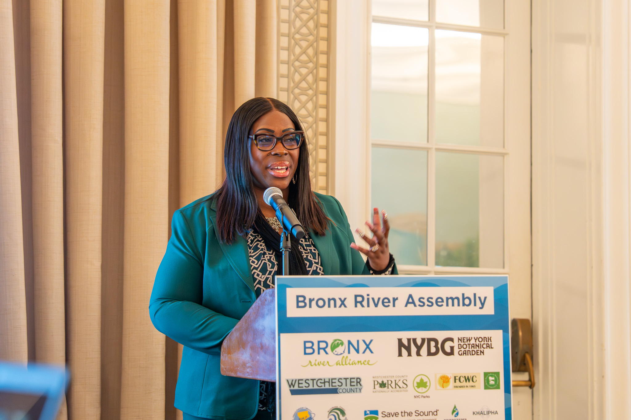 A woman with glasses, long dark hair, and a green blazer speaks at a podium.