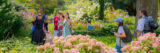 A group of students and an instructor in a summer garden full of pink and white flowers, stopping to point at a plant and make observations.