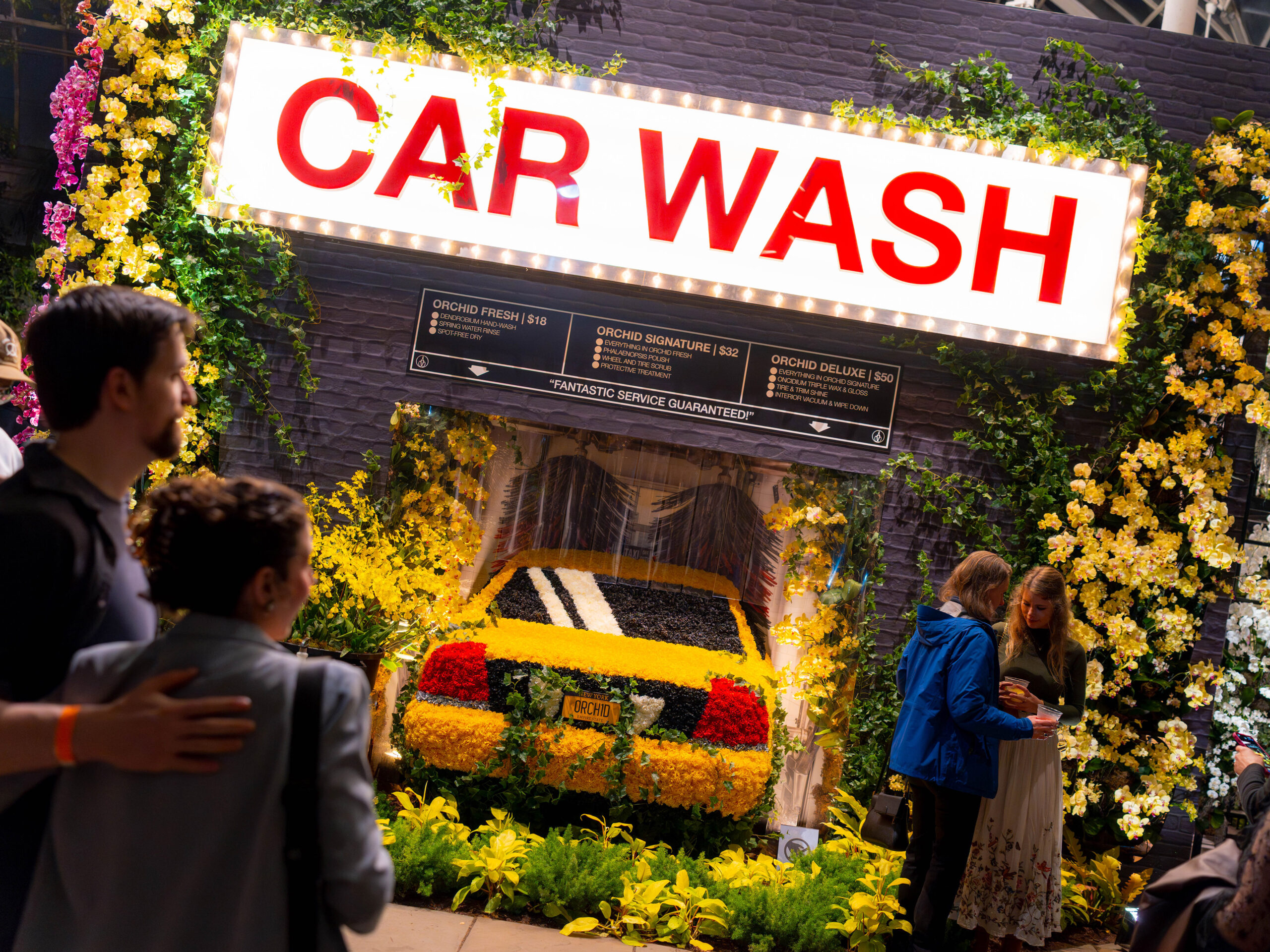 People explore a conservatory scene after dark, featuring a floral "car wash" display surrounded by flowers and a yellow cab