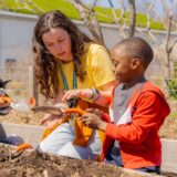 A child in an orange jacket holds a trowel up to a Camp instructor in a yellow shirt, who points to the soil and instructs the child on how to use the trowel.