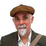 A person in a brown jacket and hat, with a white beard, poses for a photo against a white background