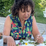 A person in a blue and green floral vest illustrates flowers in a notebook