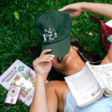 A person in a blue tank top lies on green grass, reading a white-covered paperback book while holding a green baseball cap over their face