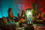 A band sits around a living room lit by a colorful pillar lamp on a table