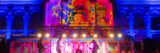 A band performs outdoors under a rainbow of colorful projector lighting