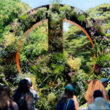 A group of people gather around a large botanical display forming the shape of a peace sign