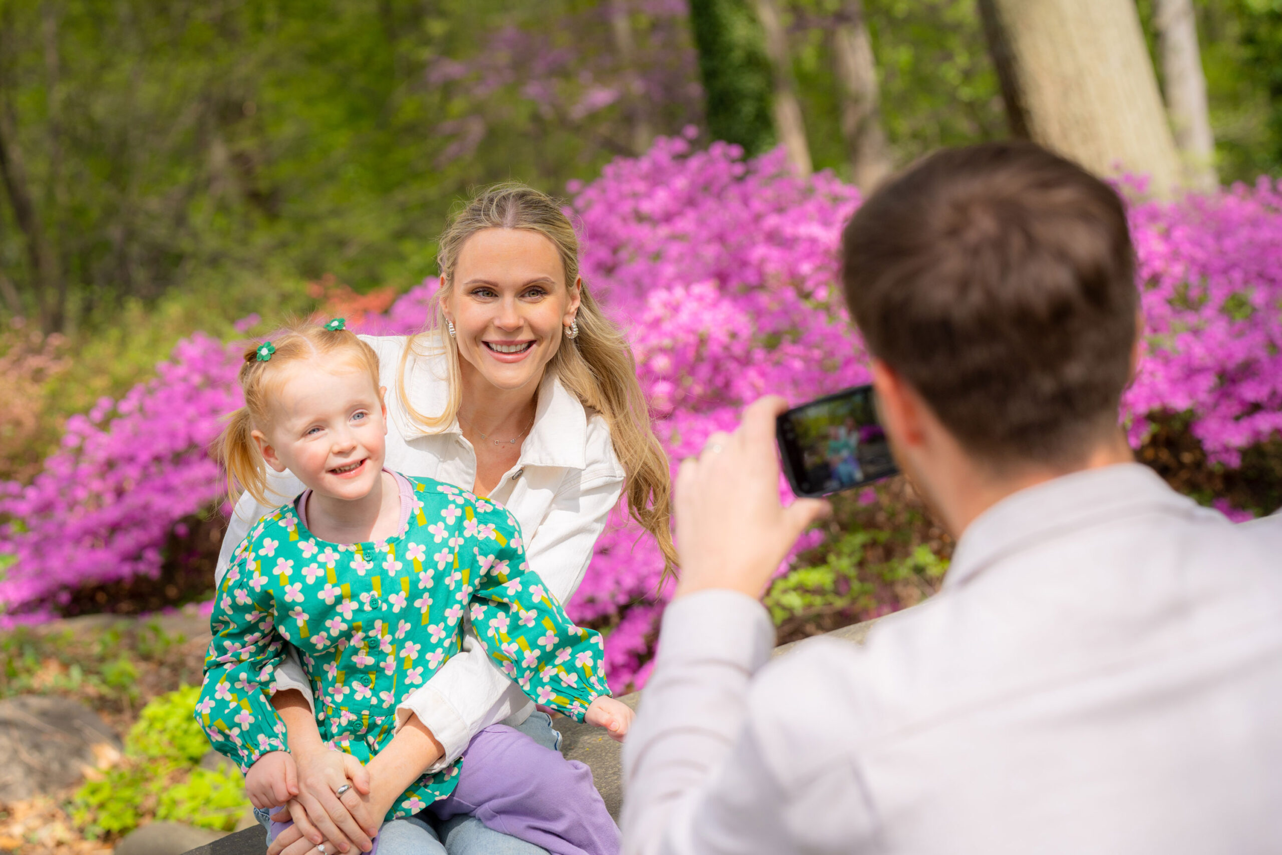 A parent and child pose for a photo in front of a sunny display of pink flowers