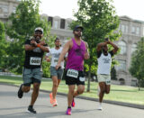 Four runners jog by green trees on a sunny day