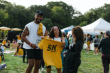 Three people hold up a yellow T-shirt with pride among a field of gathered runners