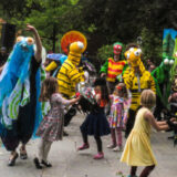 A group of colorfully dressed characters dance with children in a garden setting