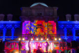 A band plays on a colorfully lit stage beneath the front facade of a large, domed building
