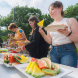 Three people fill their plates at an outdoor buffet of fruits and vegetables
