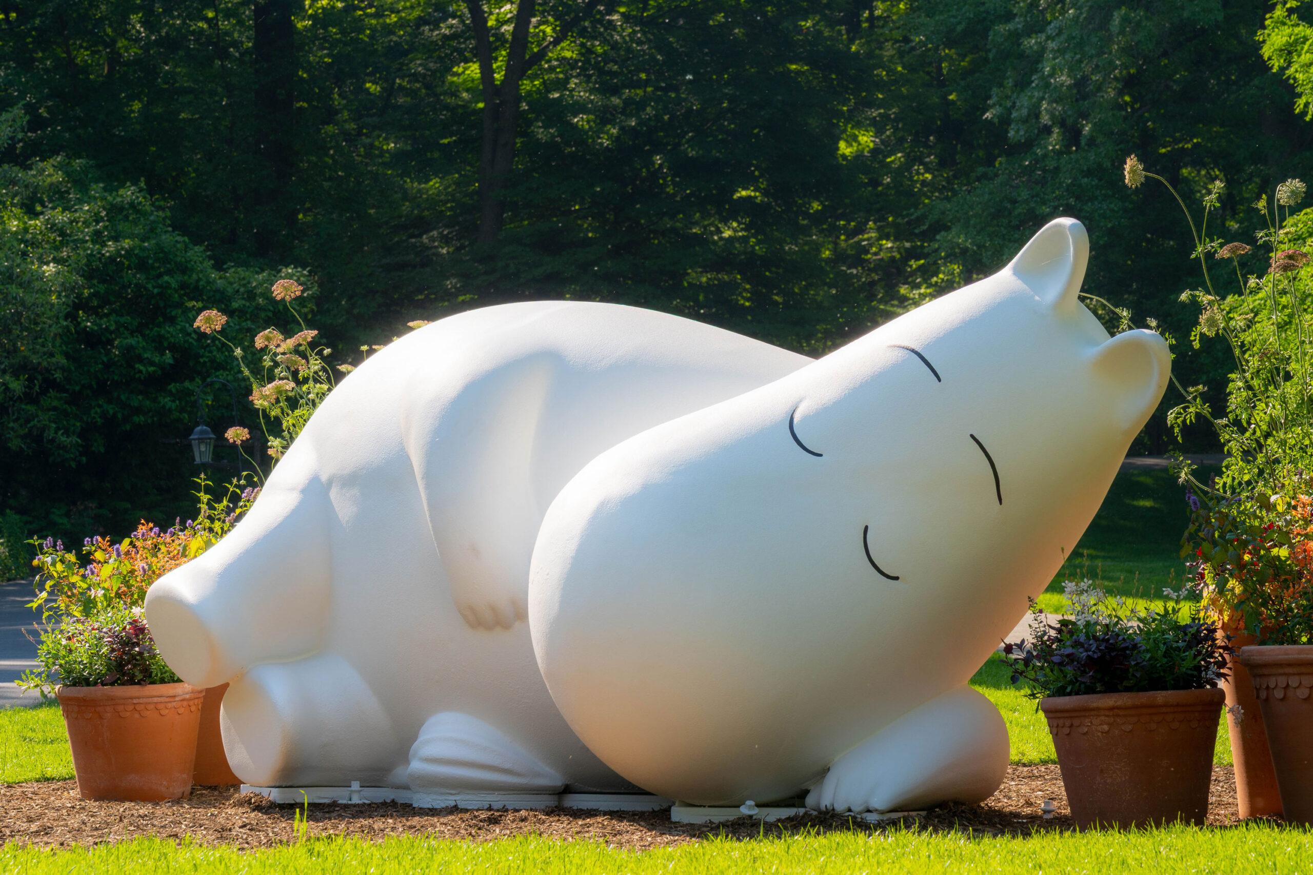 A large, white, sleeping creature statue rests on grass under the sun