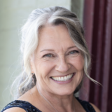 A person with long gray hair poses for a photo in a sunny room