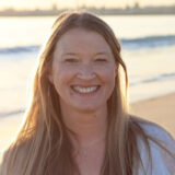 A person with long blonde hair poses for a photo on a beach at sunset