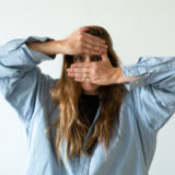 A person wearing a blue shirt, with long brown hair, poses for a photo while covering most of their face with their hands