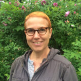 A person with short red hair and glasses poses for a photo in front of green foliage and pink flowers